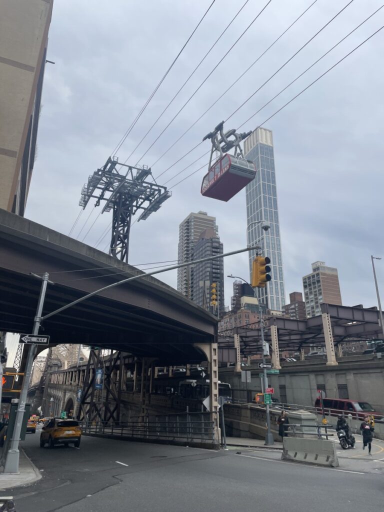 Taken near the Queensboro Bridge in Manhattan by Angela Alvarez, 2023. A red tramway heading to Roosevelt Island glides along the lines above the bridge, on a cloudy day.