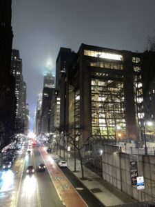 A shot on a footpath in Manhattan, looking over the fence onto the busy street with cars driving. Dimly lit buildings line the foggy streets. Shot by Angela Alvarez.