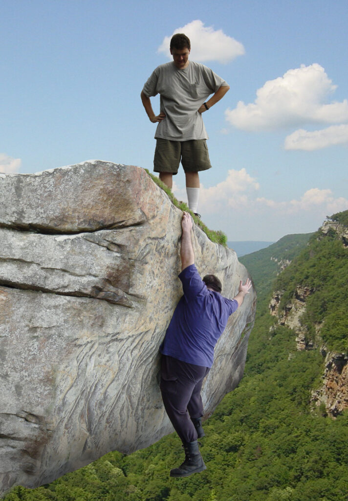One person looks on as another person hangs off the edge of a cliff.