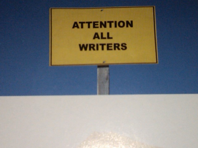 Yellow sign with black lettering that reads " ATTENTION ALL WRITERS" 