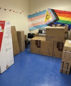 This is an image of the new Pride hang-out room at City Tech. The picture shows a blue tiled floor with a lot of boxes scattered around the room. Two flags are hanging up on the wall: a Transgender flag and an LGBTQ+ flag. The room has not been fully decorated, as the boxes in the photo contain all of the room's decorations. 