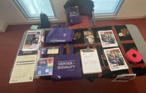 An image of a decorated table filled with Gender and Sexuality promotional fliers, stickers, tote bags, books, and raffle tickets. 