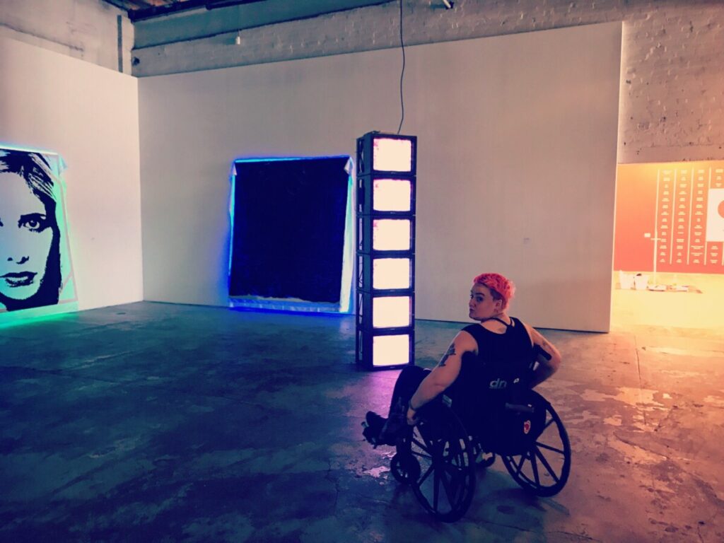 Ambient-lit photo of JRE, a white fat femme, in a manual wheelchair with bright orange hair. The floor is concrete and reflects different shades of light from the large room.