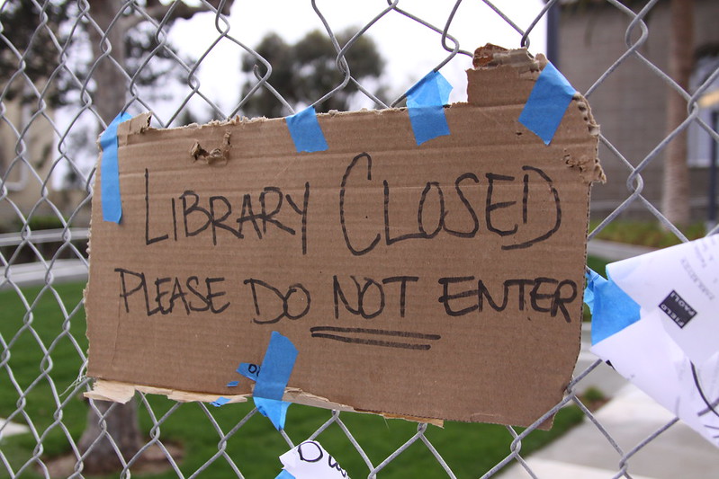 Image of a cardboard sign written in DIY sharpie style marker, stating "Library Closed Please do not enter" with the not underlined. The sign is posted using blue painter's tape against a fence in what looks like an outdoor area. 