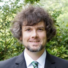 Headshot of a white man with voluminous hair and a beard smiling while wearing a black jacket and green striped tie