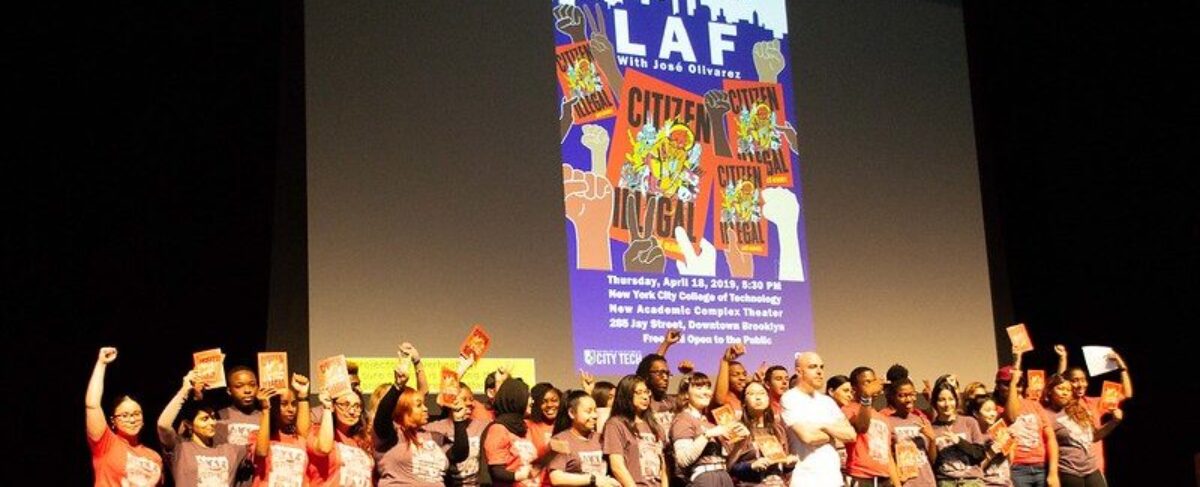 Wide-frame photo of 2019 Literary Arts Festival stage full of students and crowd members cheering and celebrating. Behind them, a large LAF poster is projected across the back of the stage.