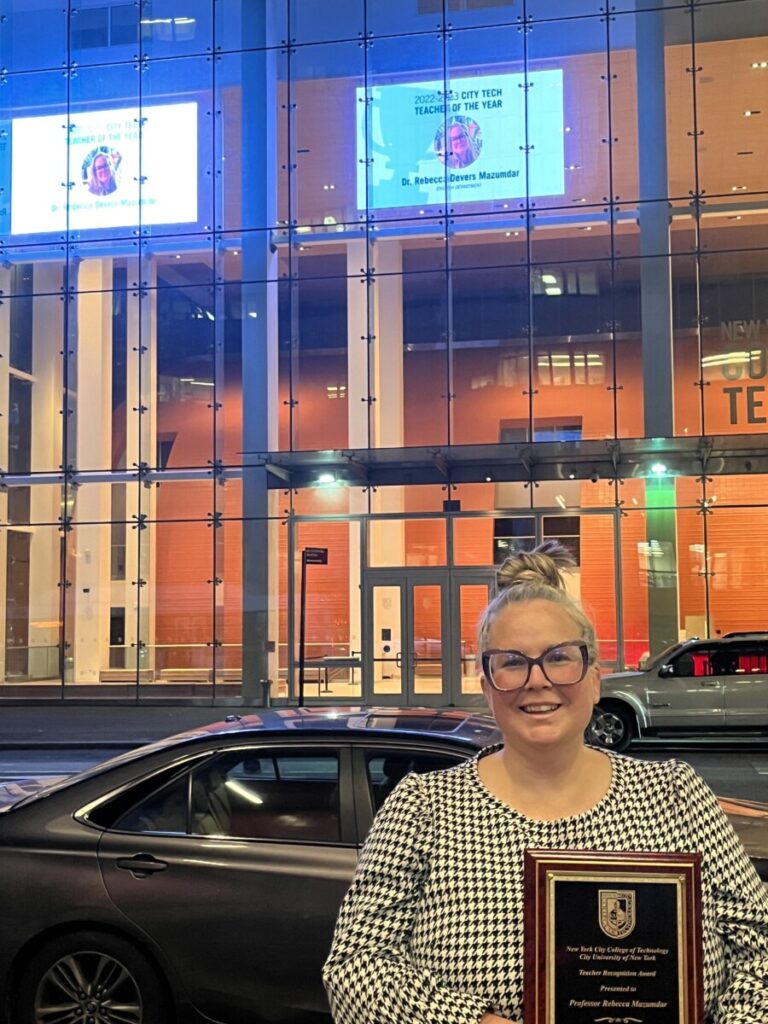 I'm pictured holding my award plaque in front of the Academic Building at City Tech. The jumbo screens on the building behind me show my photo and my name under the headline 2022-2023 City Tech Teacher of the Year.
