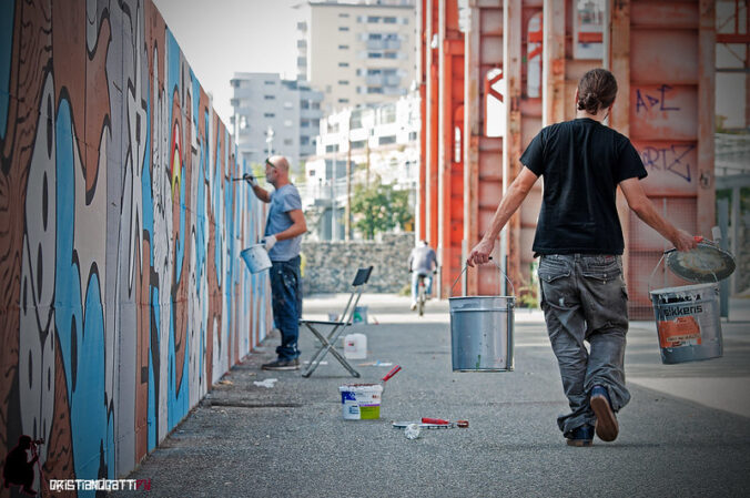 A person paints a mural outside while another person carries a bucket.