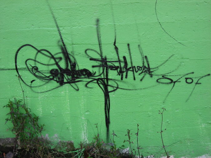 Black indecipherable graffiti on a green wall with weeds growing at the base of the wall