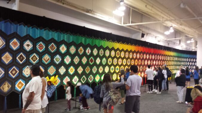 People looking at a large multicolored quilt displayed as a mural.