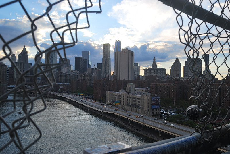 a city skyline and river's edge, as seen through a hole in a wire mesh fence